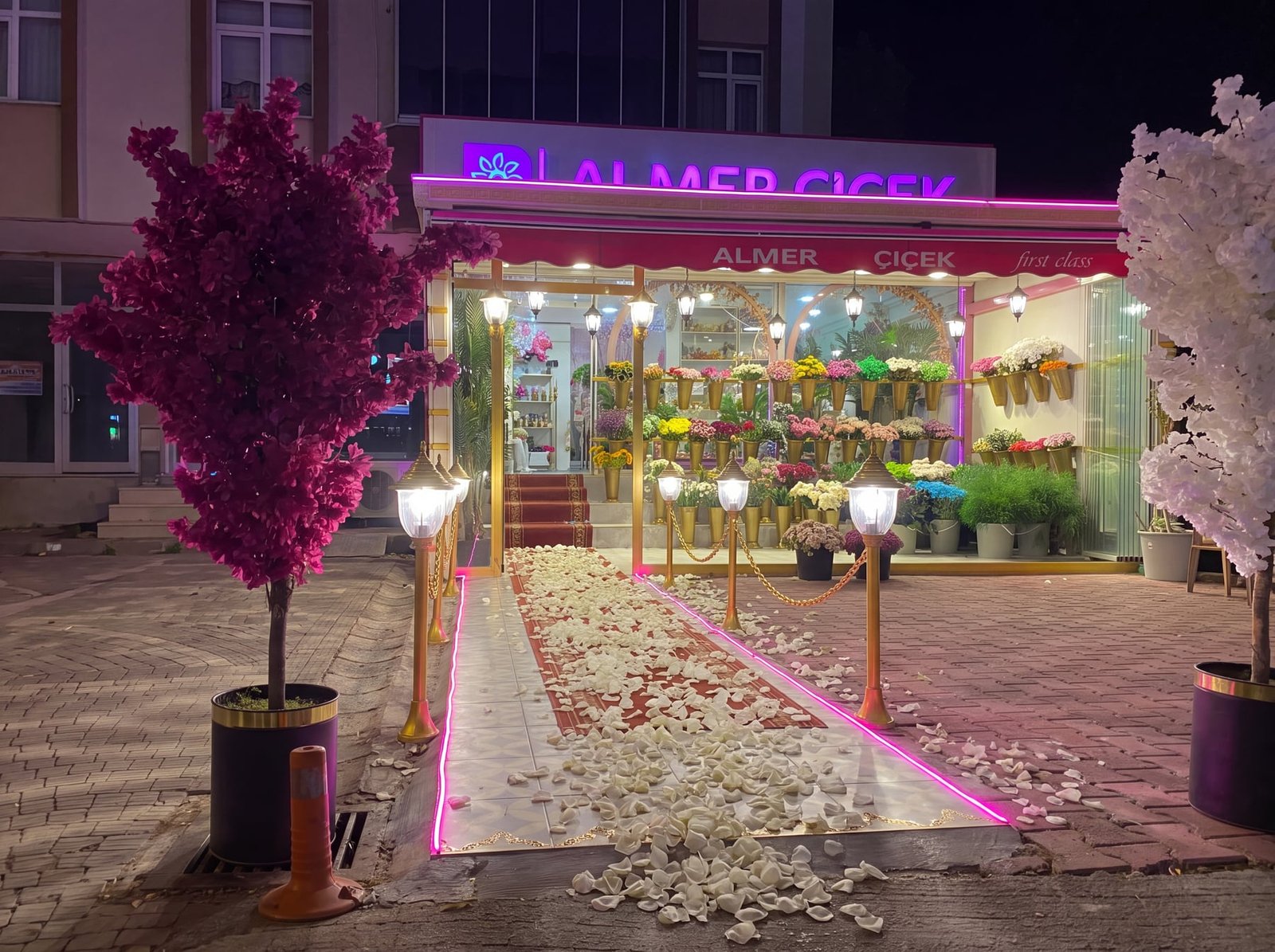 Almer Çiçek mağaza vitrini — gece ışıklandırması, taze çiçek aranjmanları ve gül yaprağı serili davet yolu